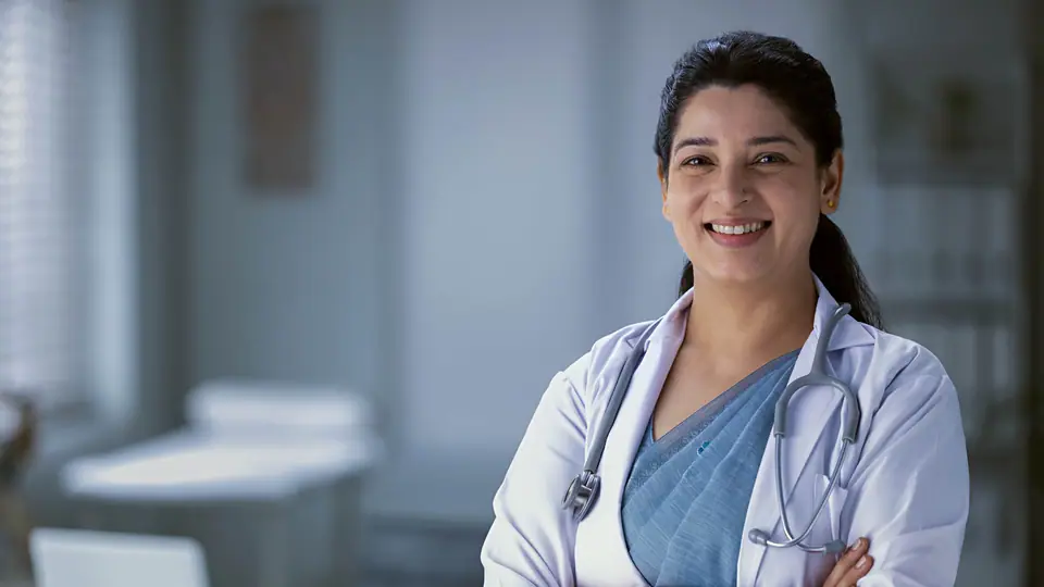 A smiling nurse in a white coat, stethoscope round her neck, arms folded