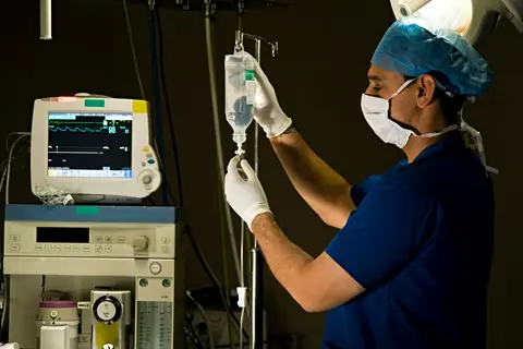 A technologist in blue scrubs and a face mask preparing an IV drip beside an anaesthesia machine