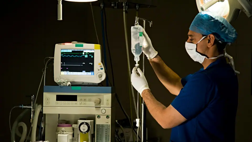 A technologist in blue scrubs and a face mask preparing an IV drip beside an anaesthesia machine