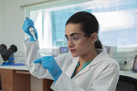 A technologist in a white coat and safety glasses pipetting a sample into a test tube in a laboratory