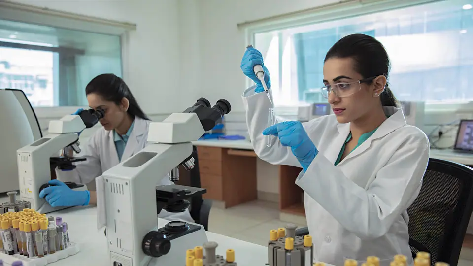A technologist in a white coat and safety glasses pipetting a sample into a test tube in a laboratory