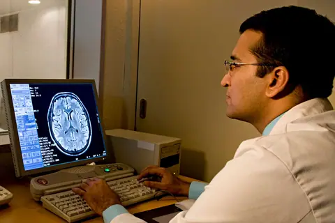A radiologist in a white coat reading a brain scan on a monitor, with a patient lying in the scanner room behind the glass