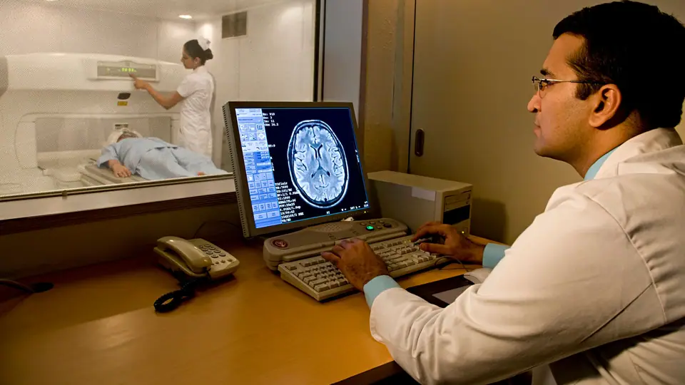 A radiologist in a white coat reading a brain scan on a monitor, with a patient lying in the scanner room behind the glass