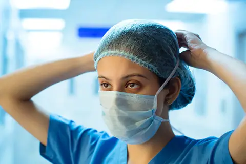 A woman in blue scrubs and a face mask adjusting her surgical cap