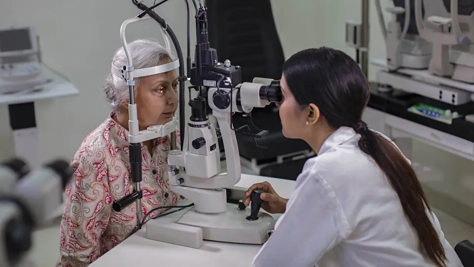 An optometrist at a slit lamp examining an older woman's eye in a clinic