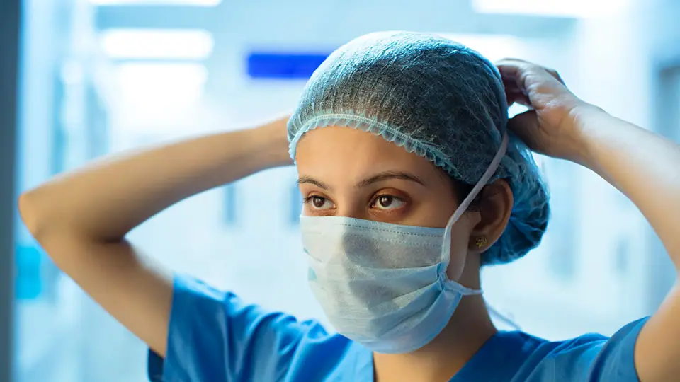 A woman in blue scrubs and a face mask adjusting her surgical cap