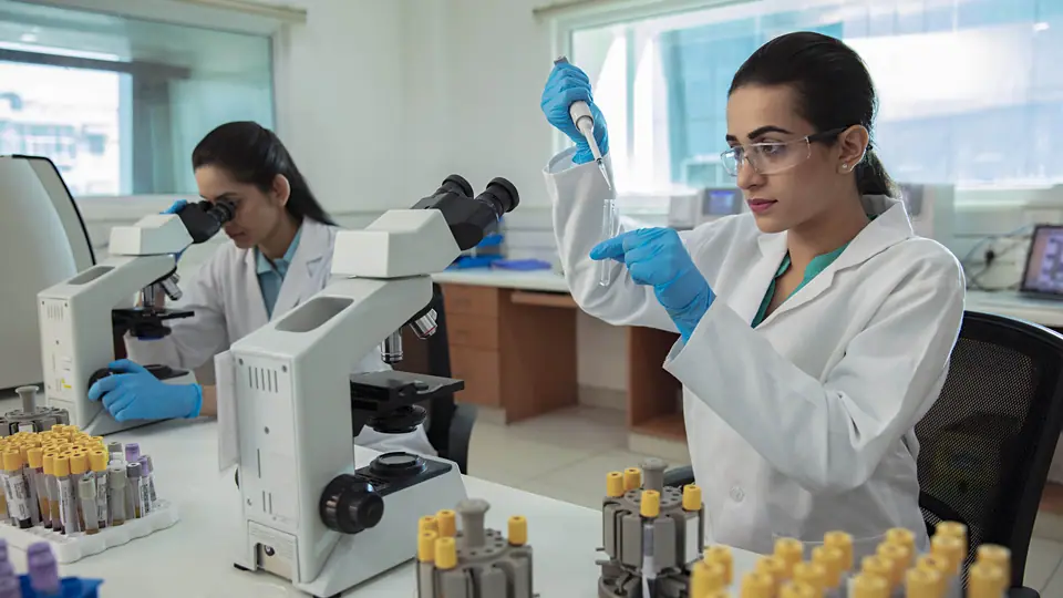 A student pipetting a sample into a test tube in a laboratory, another at a microscope