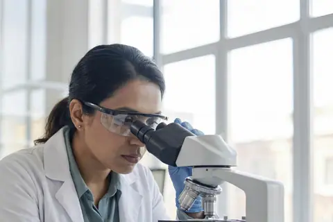 A pharmacist in safety glasses looking through a microscope in a laboratory